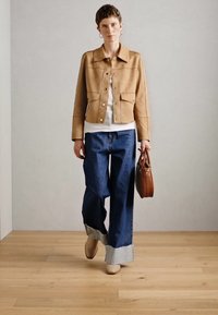 Brun mockaimitation jacka, vit topp, vida mörk denimjeans med uppvik, beiga skor, håller en brun handväska, står på trägolv.