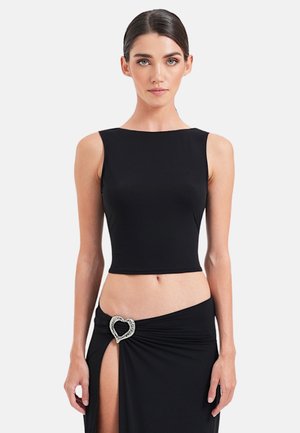 Kobieta w czarnym, bezrękawnym krótkim topie i czarnej spódnicy z rozcięciem do uda, zapinanej na ozdobną klamrę w kształcie serca z kryształkami.