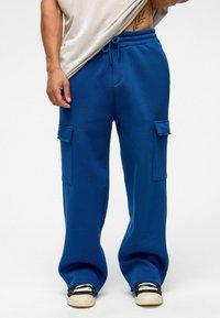 Blauwe katoenen sweatpants met een relaxte pasvorm, een trekkoord in de taille en twee zijvakjes. Gestyled met zwarte sneakers en een beige bovenstuk.
