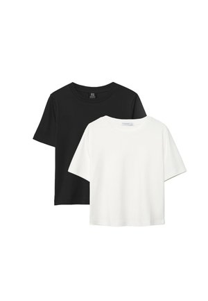 Femme aux cheveux bouclés portant un t-shirt noir à manches courtes, un short en jean clair et des sandales noires, debout les mains dans les poches.