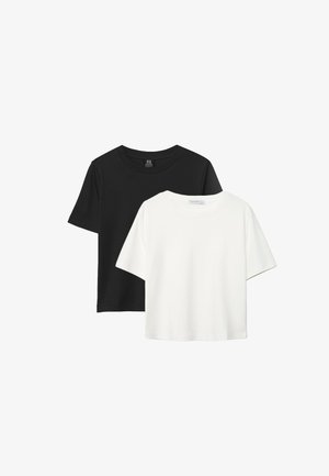 Femme aux cheveux bouclés portant un t-shirt noir à manches courtes, un short en jean clair et des sandales noires, debout les mains dans les poches.
