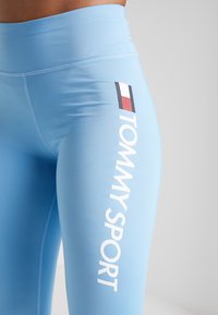 Blauwe sportleggings van rekbare stof met een hoge tailleband, witte "TOMMY SPORT" tekst en een klein logo in rood, wit en blauw.