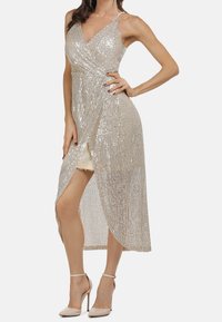 Femme en robe enveloppante sans manches en sequins argentés avec une fente, posant avec une main sur la hanche et portant des talons hauts beiges.