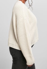 Sido vy av en person som bär en tjock krämfärgad stickad tröja med långa ärmar och mörka åtsittande jeans mot en enfärgad bakgrund.