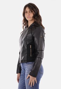 Giacca in pelle nera con cappuccio, chiusura con zip davanti e tasche laterali. Presenta un design aderente e pannelli in tessuto a contrasto. Accentata da hardware dorato.