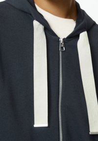 Felpa con zip di colore blu scuro realizzata in un tessuto testurizzato. Presenta cordini bianchi e una cerniera argentata, con un colletto a costine sotto.