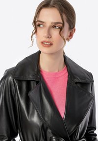 Zwarte leren trenchcoat met een brede kraag, een ceintuur in de taille en een glanzende textuur, gelaagd over een roze trui.