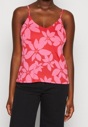 Femme portant un débardeur rouge avec de grands motifs floraux roses et un pantalon noir, debout devant un fond clair uni.