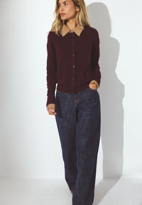 Bordový kardigán s béžovým golierom, zapínaním na gombíky a dlhými rukávmi, skombinovaný s voľnými tmavomodrými džínsami. Hladká textúra a priliehavý strih.