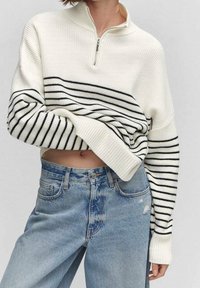 Biały dziany sweter z kołnierzem na półzamek, z czarnymi poziomymi paskami. Połączony z jasnoniebieskimi dżinsami z przetarciami na kolanie.