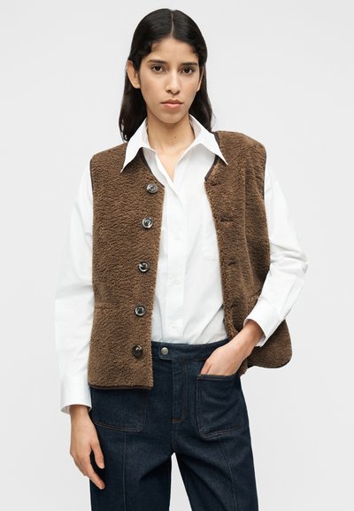 Καφέ γιλέκο από μάλλινο ύφασμα με πέντε κουμπιά, φορεμένο πάνω από λευκό πουκάμισο. Διαθέτει άνετη εφαρμογή και στρογγυλεμένες άκρες. Σχετικά σκουρόχρωμο τζιν παντελόνι φαίνεται από κάτω.