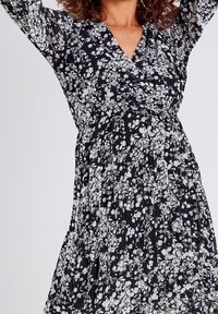 Robe noire à motifs floraux avec décolleté en V, manches longues et taille froncée. Présente des motifs de fleurs blanches et un design fluide et plissé.