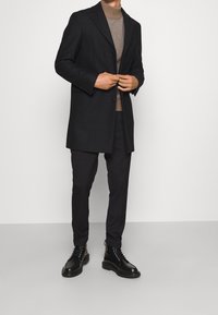 Schwarzer Wollmantel mit einem einzelnen Knopfverschluss, getragen über einem braunen Rollkragenpullover und schwarzen Hosen; kombiniert mit schwarzen Schnür-Stiefeletten.