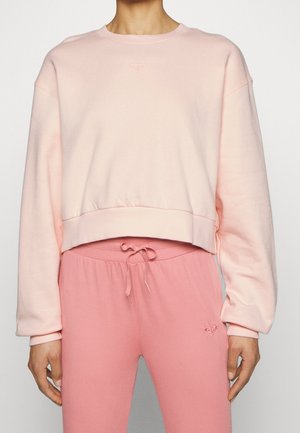 Person iført lyserød cropped sweatshirt og matchende lyserøde sweatpants med snøre og små hjerte-logoer på bryst og lår.