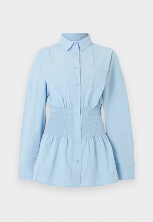 Vrouw draagt een lichtblauw oversized overhemd met knopen, getailleerd met een ceintuur, zwarte minirok met riem, zwarte laarzen tot halverwege de kuit en donkere zonnebril.