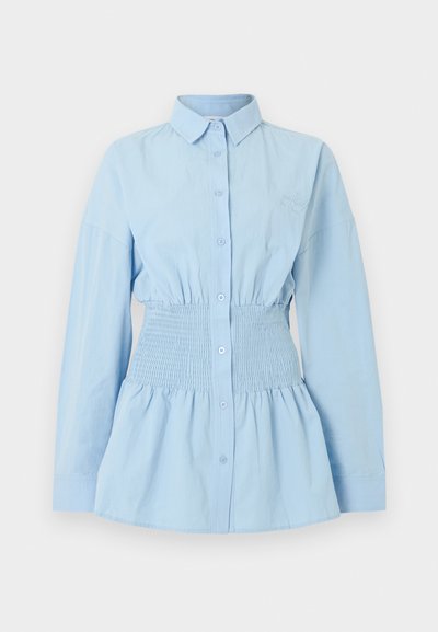 Ανοιχτό μπλε μπλούζα με μακριά μανίκια, κουμπιά στο μπροστινό μέρος, ελαστική μέση με σούρα και γιακάς τύπου spread σε απλό φόντο.