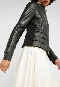 Black leather jacket with layered texture on sleeves and fitted design. Features a high collar and silver zipper, paired with a light-colored skirt.