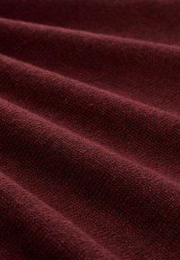 Burgunderroter Stoff mit strukturierter Oberfläche, der sanfte, fließende Falten zeigt. Die Webart ist dicht, was seine Wärme und haptische Qualität verbessert.