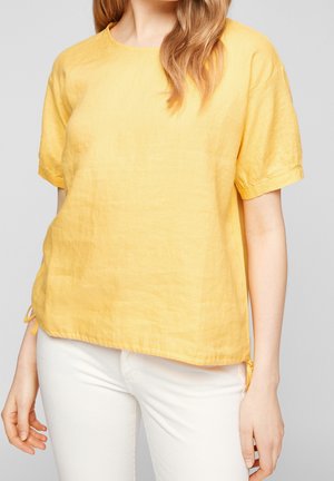 Vrouw draagt een losse, geelgekleurde blouse met korte mouwen en bindstrikjes aan de zoom, gecombineerd met witte, nauwsluitende broek, tegen een grijze achtergrond.