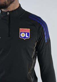 Chaqueta deportiva negra con cuello alto, cierre de cremallera y franjas de acento azules. Muestra el logo del Olympique Lyonnais en la parte frontal.