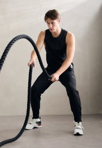 Zwarte sportieve tanktop en gestructureerde zwartebroek, terwijl ze een dikke zwarte battle rope vasthoudt. Schoenen zijn wit met zwarte en rode accenten. Neutrale achtergrond.