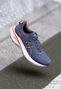 Gray athletic shoe with orange and black accents, lightweight design, textured upper, cushioned sole, and logo on the side.