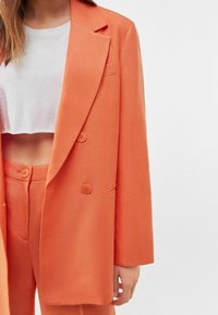 Oranžový dvouřadý blazer s klopami, předními kapsami a texturovaným materiálem. Nesený přes bílý krátký top.