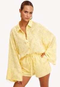 Chemise jaune texturée avec manches longues évasées, boutonnée à l'avant, assortie à un short. Tissu doux avec détails effilochés.