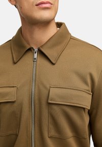 Brun glidelåsjakke laget av teksturert stoff. Har krage og to fremre klafflommer med synlige sømmer og metallglidelås.