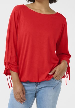 Femme portant un haut rouge à manches longues froncées et un jean bleu clair, une main dans la poche, sur un fond uni.