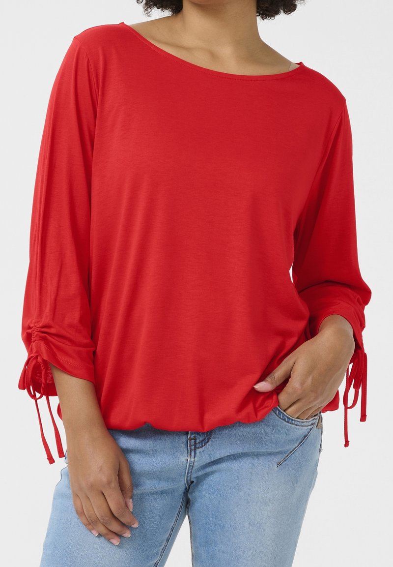 Femme portant un haut rouge à manches longues froncées et un jean bleu clair, une main dans la poche, sur un fond uni.