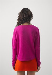 Fuchsia gestrickter Pullover mit langen Ärmeln und rundem Ausschnitt, kombiniert mit einem orangefarbenen Miniröckchen. Der Rückansicht zeigt eine lockere Passform und gerippte Säume.