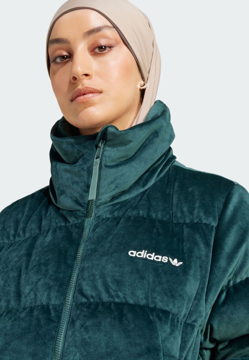Dunkelgrüne, weiche, mit Reißverschluss versehene Jacke mit hohem Kragen und Adidas-Logo. Besitzt eine gesteppten Textur und ein weiches Material.