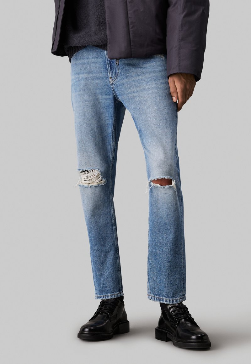 Jeans strappati di un blu chiaro con ginocchia sfrangiate, design a gamba dritta, abbinati a scarpe nere con lacci. Denim testurizzato con un aspetto usurato.