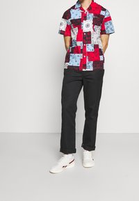 Maglietta a maniche corte con design patchwork; i colori includono rosso, blu e bianco. Indossata con pantaloni neri e sneaker bianche con accenti rossi.
