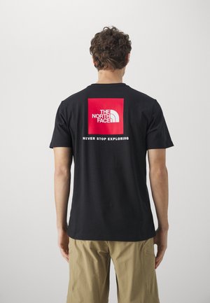 Homme de dos portant un t-shirt noir avec un logo carré rouge et le texte "The North Face Never Stop Exploring" ainsi qu'un short beige.
