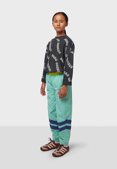 Schwarzes Langarmshirt mit grauem Text, kombiniert mit hellgrünen Hosen mit blauen Streifen und einem gelben Kordelzug. Braune Schuhe mit Streifen.