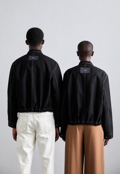 Schwarze kurz geschnittene Jacken mit kontrastierenden weißen Nähten; elastischer Bund; Etiketten auf der Rückseite. Ein Modell trägt weiße Hosen, das andere braune.