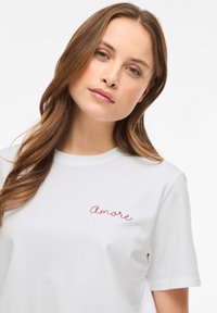 Νέα γυναίκα με μακριά καστανά μαλλιά, φορώντας λευκό T-shirt με κόκκινη κεντημένη επιγραφή «amore» στο στήθος, σε απλό λευκό φόντο.