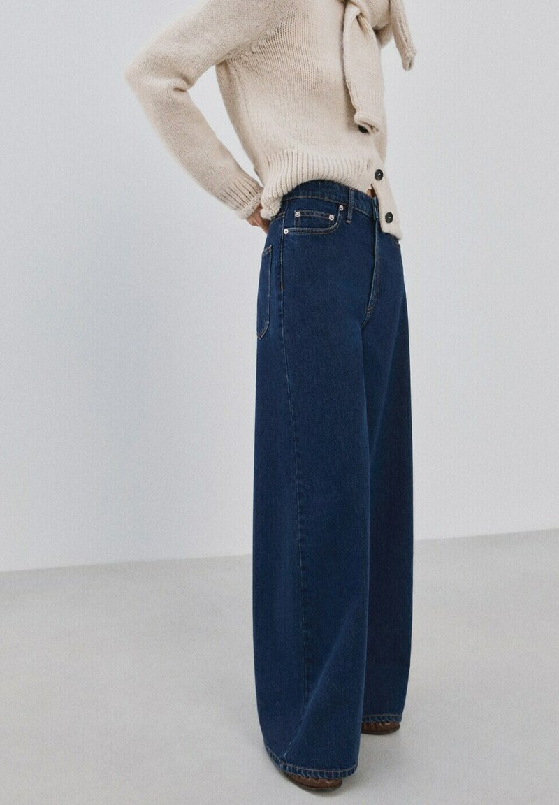 Personne portant un jean large taille haute bleu foncé et un cardigan beige en maille avec des boutons, debout les mains derrière le dos.