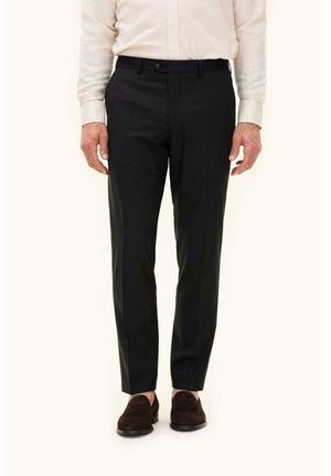 Uomo con pantaloni eleganti neri, camicia beige a maniche lunghe con bottoni e mocassini scamosciati marrone scuro, in piedi contro uno sfondo chiaro.