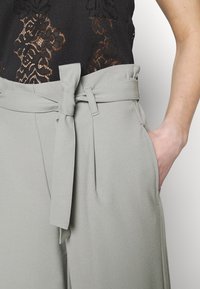 Svart spets topp parat med högmidjade grå byxor som har ett knytet tygbälte och handen i höger ficka.