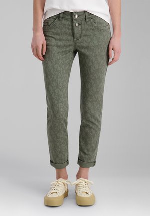Beine, die olivgrüne Slim-Fit-Hosen mit dezentem Blumenmuster tragen, mit hochgekrempelten Säumen, weißen Sneakers mit beigen Sohlen und einem weißen Oberteil.