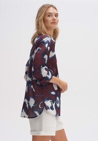 Langärmlige, kastanienfarbene Bluse mit blauem und weißem abstraktem Muster. Leichter, durchsichtiger Stoff mit seitlichem Schlitzsaum. Kombiniert mit weißen Shorts.