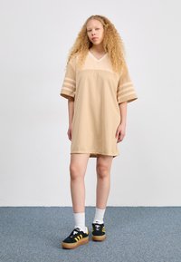 Vestido de red oversize en color beis con escote en V y mangas cortas con rayas. Combinado con zapatillas negras y calcetines blancos sobre un fondo de moqueta azul.