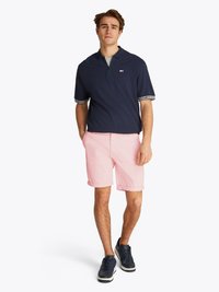 Polo azul-marinho com fecho de zíper e padrão branco em contraste nas mangas, combinado com calções cor-de-rosa claros e ténis azul-marinho.