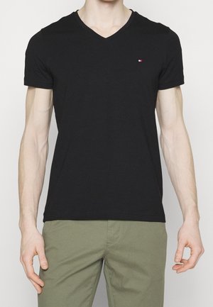 Homme portant un t-shirt noir ajusté à col en V avec un petit logo rouge, blanc et bleu sur la poitrine gauche et un pantalon vert olive.