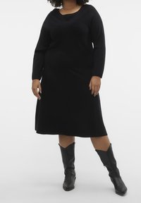 Robe noire en tricot à manches longues avec un décolleté en scoop, une silhouette fluide et une texture douce, portée avec de hautes bottes noires de style cowboy.