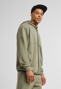 Khaki bluza z kapturem na zamek, z kieszeniami po bokach i ściągaczami na rękawach. Wykonana z miękkiego materiału, o minimalistycznym designie.