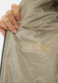 Grijze, gladde stofinterieur met waterafstotende, winddichte en donsvrije iconen in oranje gemarkeerd. Zichtbare ritssluiting.
